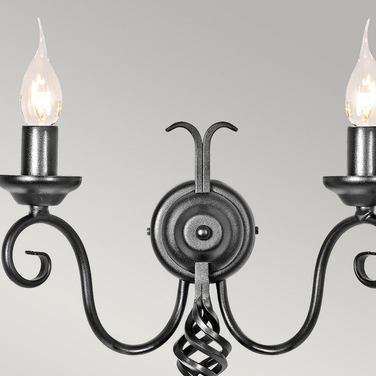 The Harlech 2 Light Wall Light in black displays a medieval-inspired design with its candelabra-style arms and a twisted central column, boasting two candle-shaped light bulbs. Set against a light gray background, it exudes timeless elegance and sophistication with its sleek wrought iron construction.