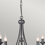 A close-up of the Harlech 5 Light Chandelier - Black features lit candle-style bulbs, with a black metal chain that enhances its rustic charm against a plain, light-colored wall. This chandelier exudes an elegant yet timeless ambiance.