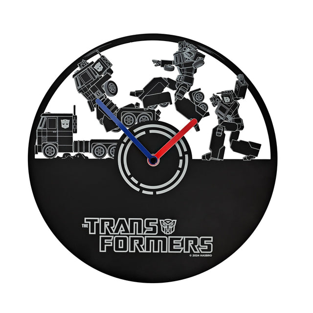 Round black metal wall clock, 30cm, with cut-out scene of a truck transforming into a robot in fine grey lines, blue and red hands, "The Transformers" logo on the plain lower half.