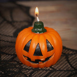 Lit Halloween Pumpkin Debossed Smiley Face Candle, 7cm round orange ribbed wax with black jack-o'-lantern face and dark green stem wick, shown on spider web decor.