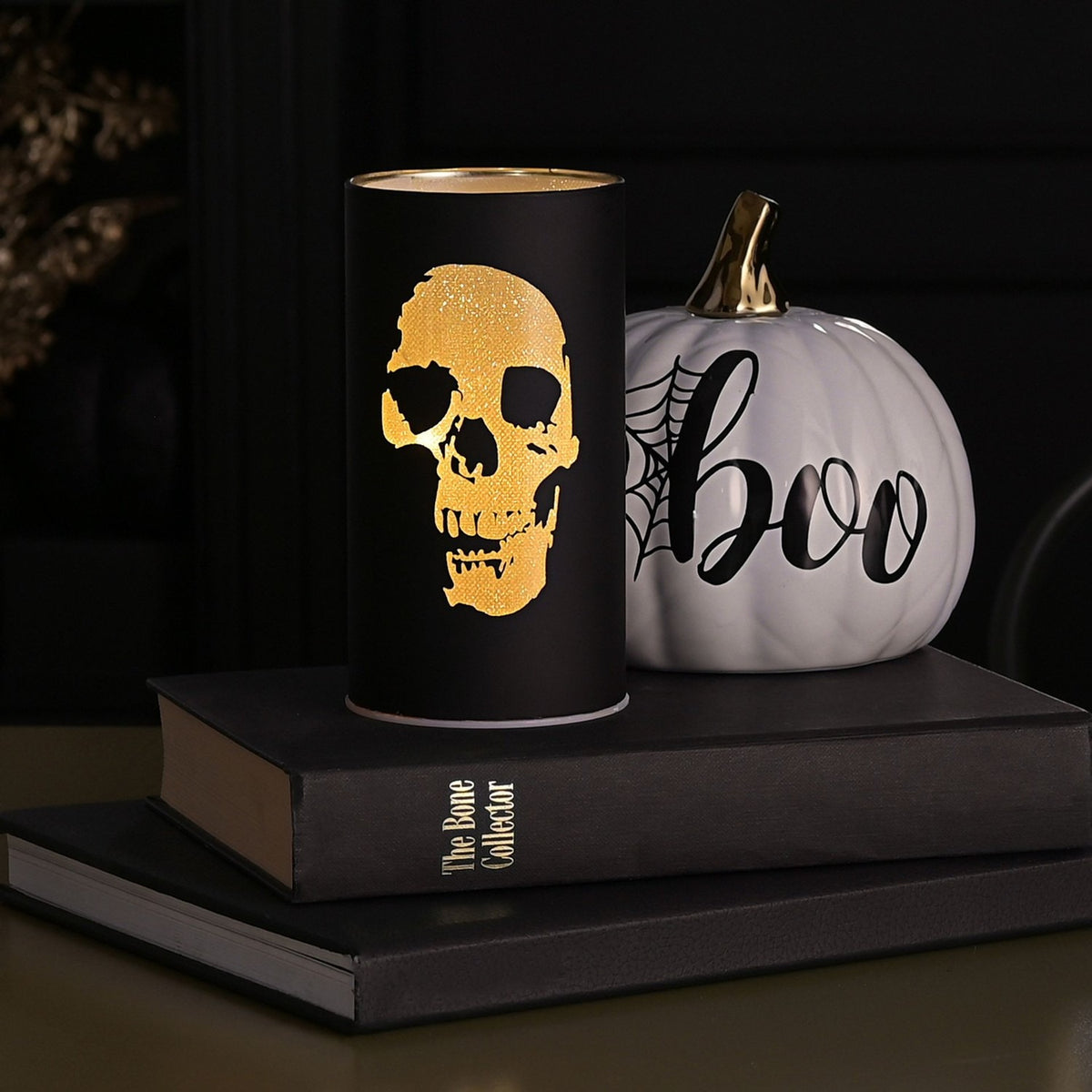Round black LED lantern, 15.2cm, with a glowing gold skull silhouette design, displayed on books beside a white "boo" pumpkin in a Halloween setting.