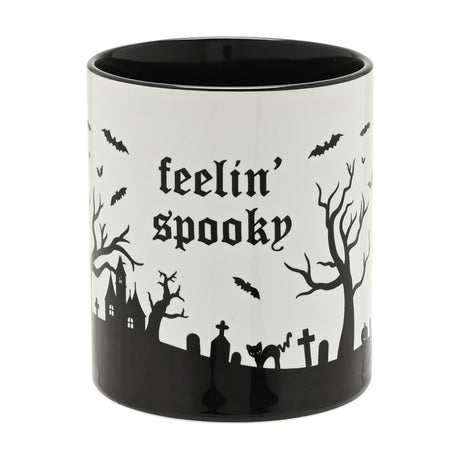 White glazed stoneware mug reading "feelin' spooky" in black gothic text, with a silhouetted graveyard scene, bats, haunted house and black cat around a black base, on a plain background.
