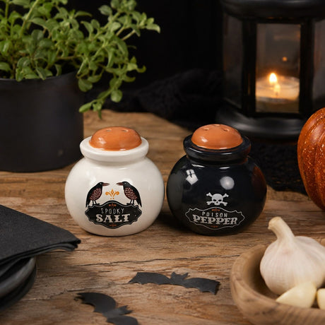 Stoneware salt and pepper set with orange domed lids: white Spooky Salt shaker with crows and black Poison Pepper shaker with skull and crossbones, on a wooden table with Halloween decor.