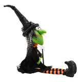 Small sitting witch Halloween ornament with a lime-green face, tall bent black hat, black dress with orange buttons and bow, striped stockings, and star wand, shown on a plain white background.