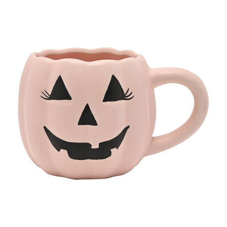 Pink pumpkin-shaped mug with scalloped rim and ribbed body, featuring a black jack-o'-lantern face with triangle eyelashed eyes and a gap-toothed smile, set on a plain white background.