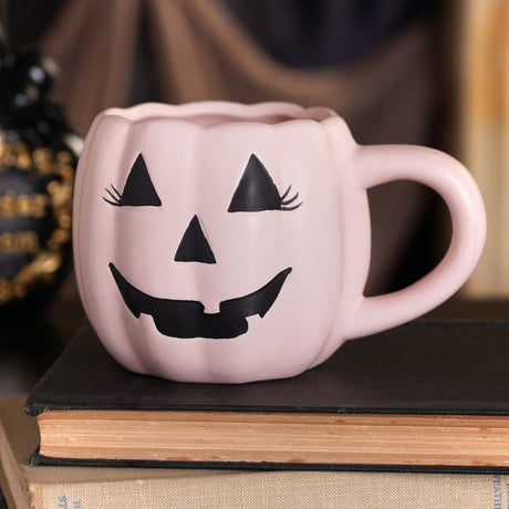 Pink pumpkin-shaped mug with ribbed body, scalloped rim, and black jack-o'-lantern face with eyelashes, displayed on a stacked book against a dark backdrop.
