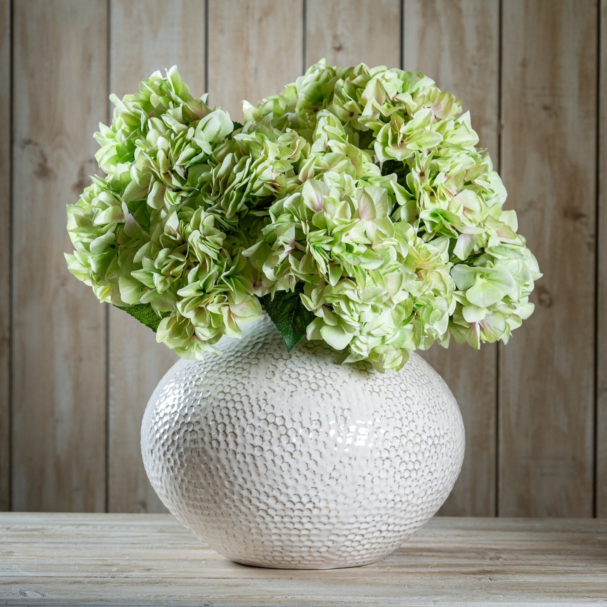 Hill Interiors Shabby Green Hydrangea Bouquet arranged in a textured white ceramic vase on a wooden surface against a pale plank background.