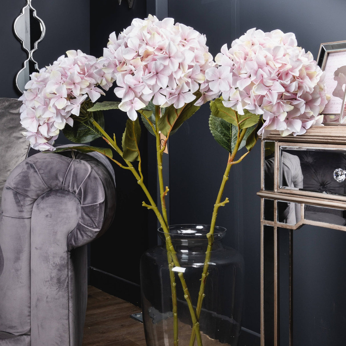Three Hill Interiors Giant Pink Hydrangea stems with full rounded blush blooms displayed in a clear glass vase beside a velvet sofa and mirrored console.