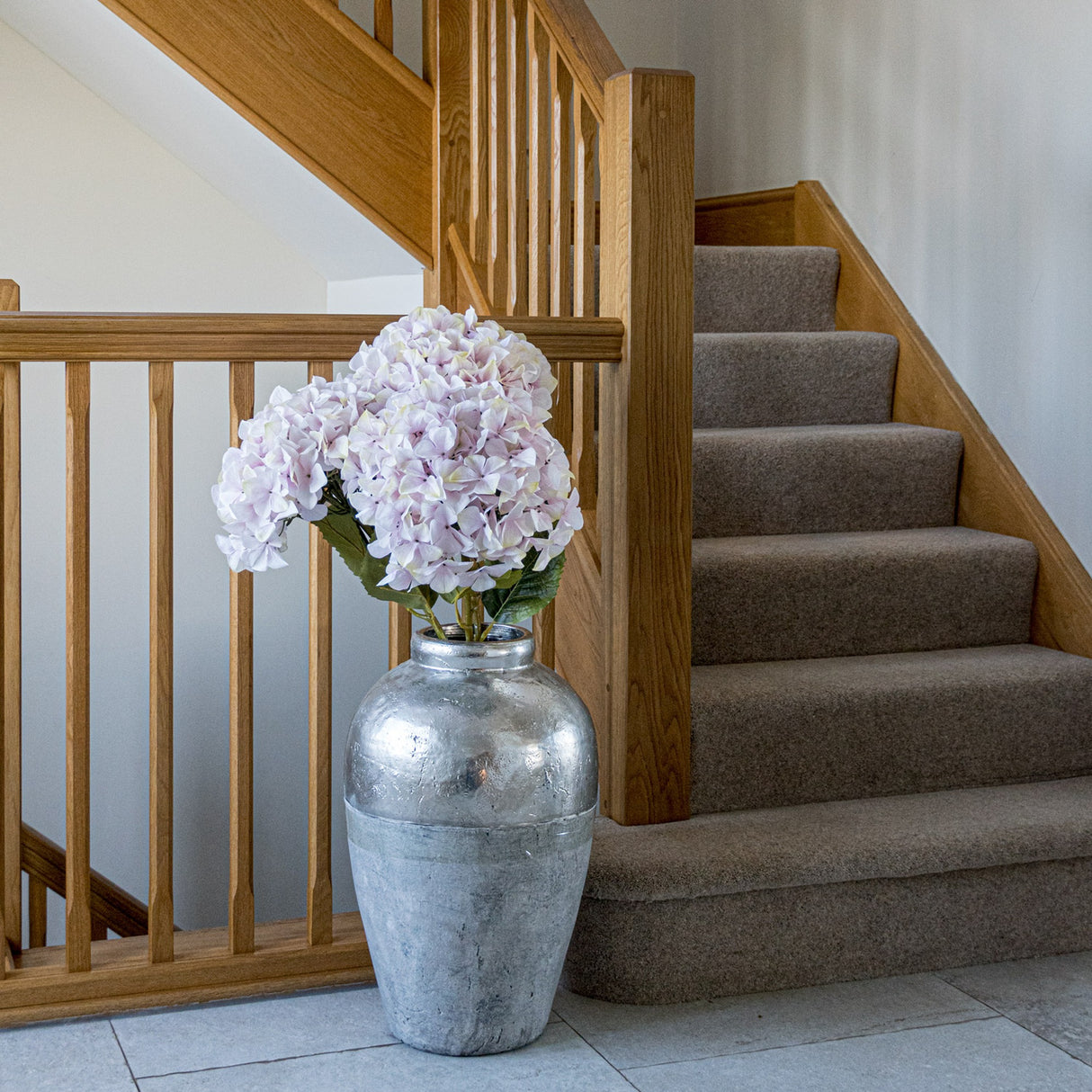 Hill Interiors Giant Pink Hydrangea faux stem with a large rounded pale pink bloom, displayed in a silver vase beside an oak staircase.