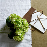 Hill Interiors Green Hydrangea Bunch of faux stems laid on white linen beside a leather-bound journal and pencil on hessian fabric.