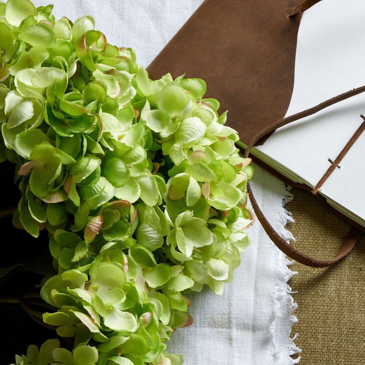 Hill Interiors green hydrangea bunch with layered florets displayed beside a brown leather-bound journal on a fringed white cloth.