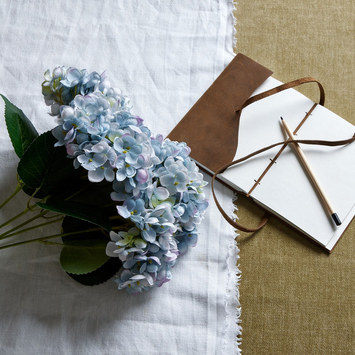 Hill Interiors Blue Hydrangea Bunch with pompom blooms and green leaves on a fringed white linen cloth beside a leather notebook and pencil.