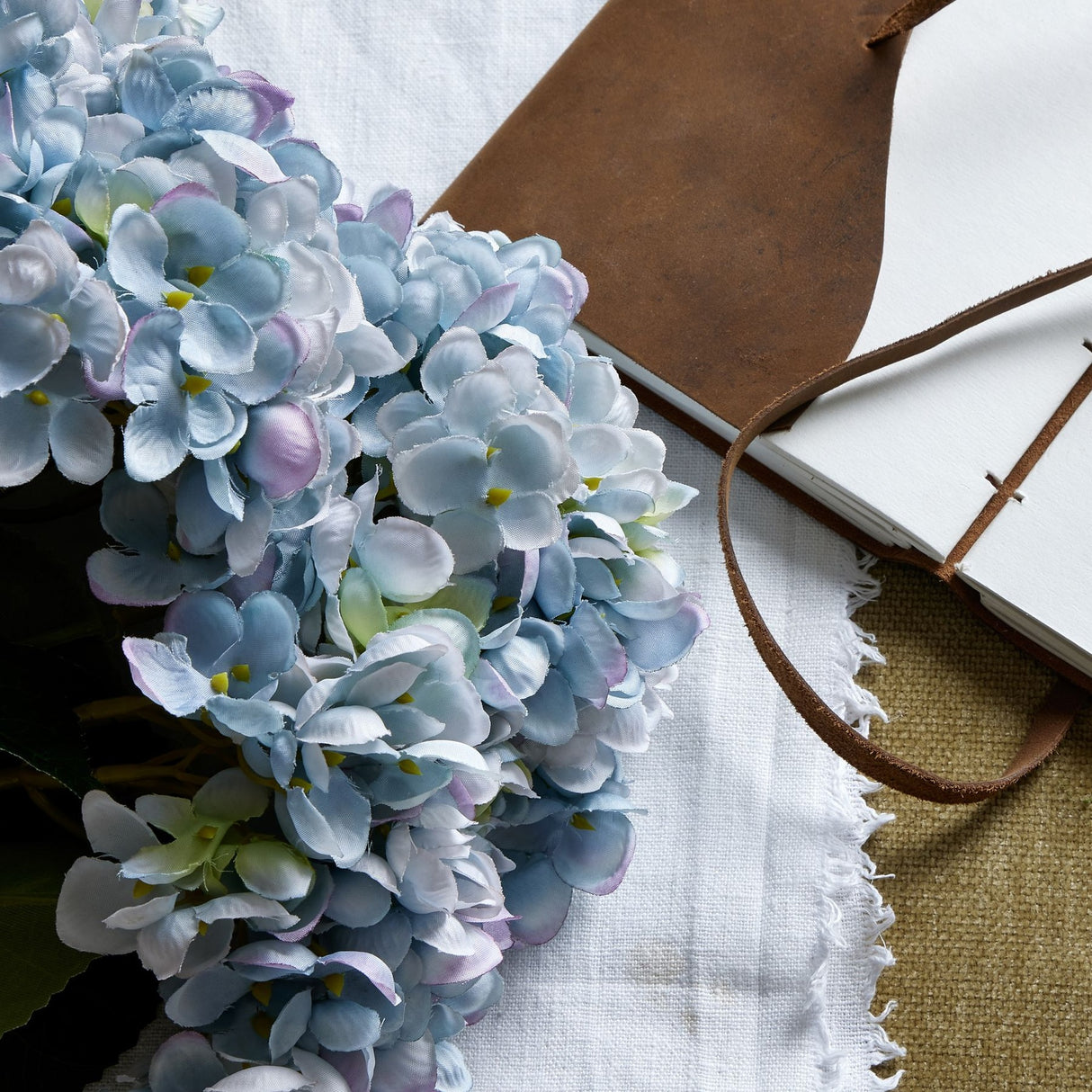 Hill Interiors Blue Hydrangea Bunch faux flowers with clustered blue florets, styled beside a leather-handled notebook on a fringed white linen cloth.