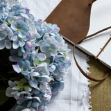 Hill Interiors Blue Hydrangea Bunch faux flowers with clustered blue florets, styled beside a leather-handled notebook on a fringed white linen cloth.