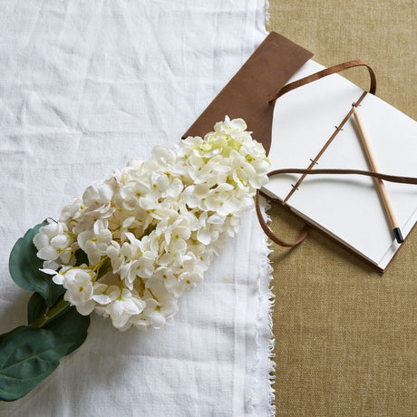Hill Interiors White Spear Hydrangea faux stem with dense white four-petal florets and green leaves, laid on white linen beside a leather journal.