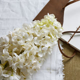 Hill Interiors White Spear Hydrangea faux stem with a full cluster of small four-petal white florets, resting on linen beside a tan leather-bound journal.