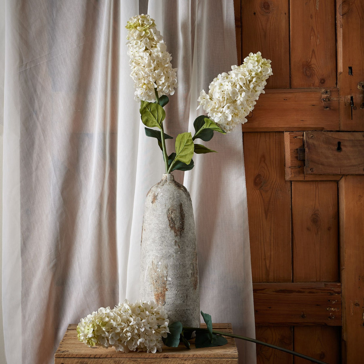 Hill Interiors White Spear Hydrangea faux stems in a tall distressed stone vase on a wooden crate beside sheer curtains and a rustic wooden door.