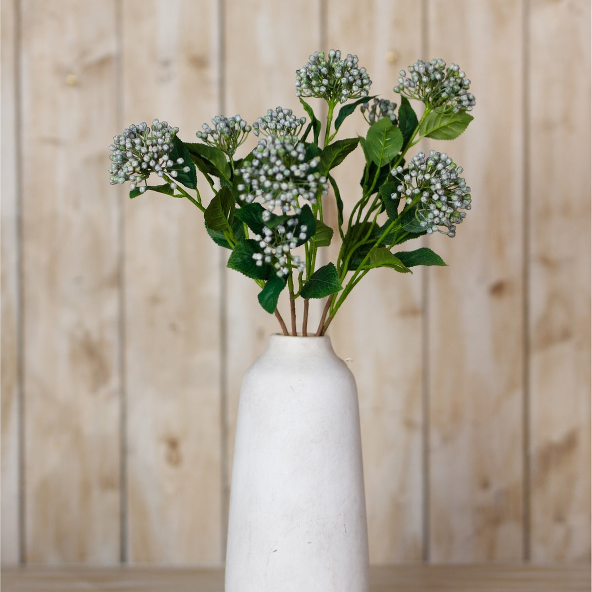 Hill Interiors Blue Budding Hydrangea Spray artificial stems with clustered blue-grey buds and green leaves arranged in a white ceramic vase against light wood.