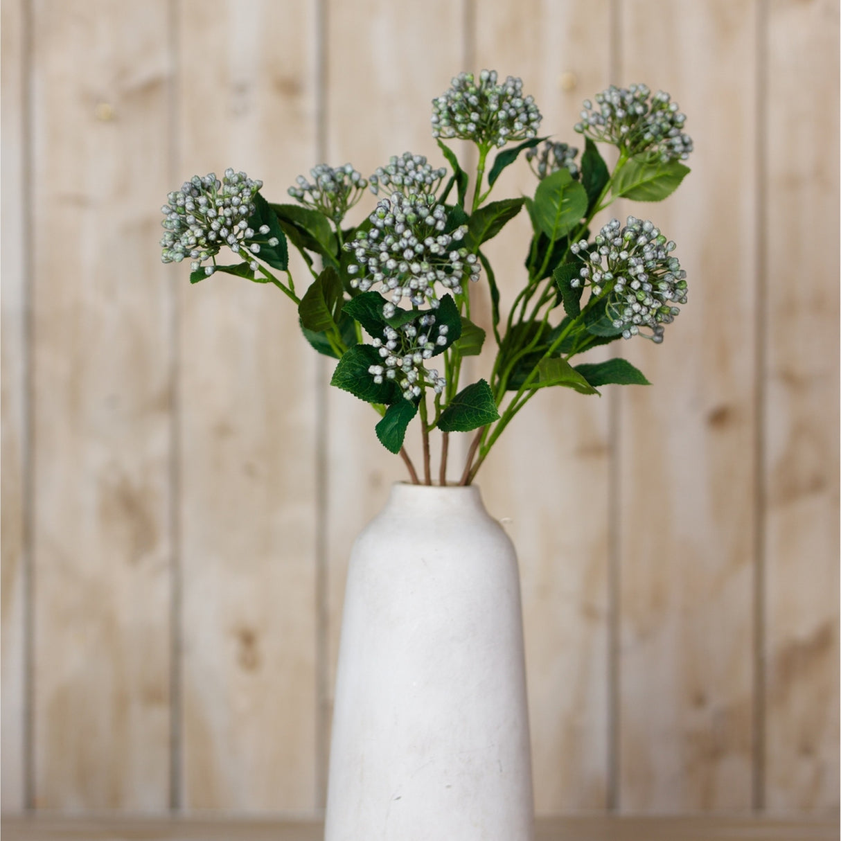 Hill Interiors Blue Budding Hydrangea Spray in a white ceramic vase against a light wood panel background, with clustered blue buds and green leaves.