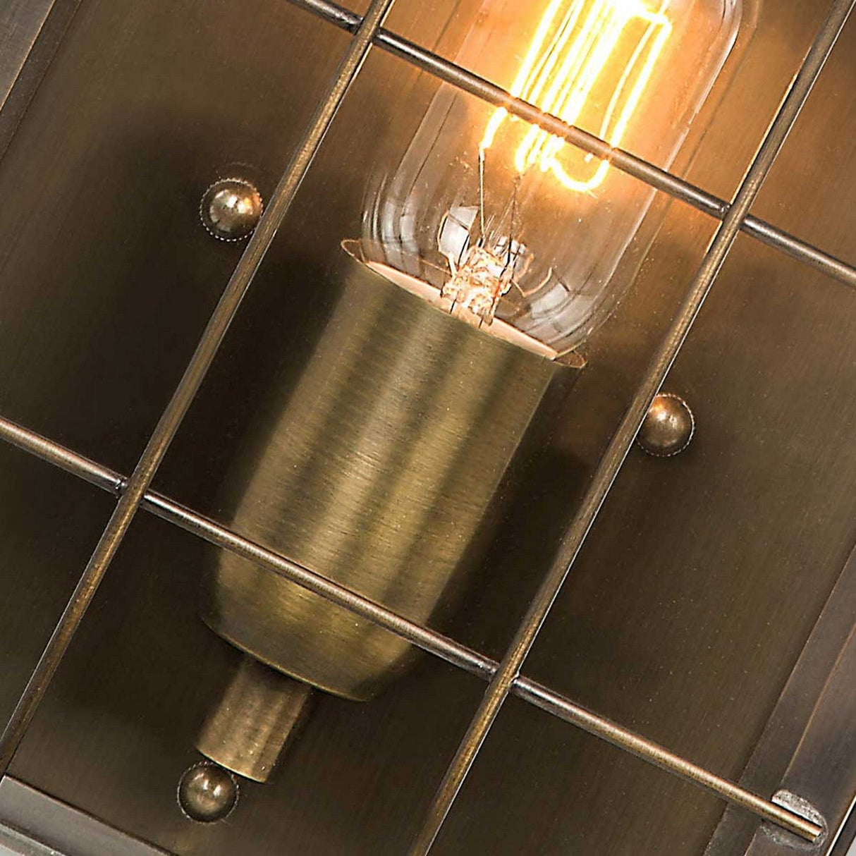 Close-up of a Hyde 1 Light Outdoor Wall Light in aged brass, featuring an illuminated vintage-style bulb encased within a metal grid frame, showcasing its retro charm. The light bulb emits a warm glow, accentuating the aged brass socket and the geometric design of the grid.