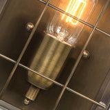 Close-up of a Hyde 1 Light Outdoor Wall Light in aged brass, featuring an illuminated vintage-style bulb encased within a metal grid frame, showcasing its retro charm. The light bulb emits a warm glow, accentuating the aged brass socket and the geometric design of the grid.