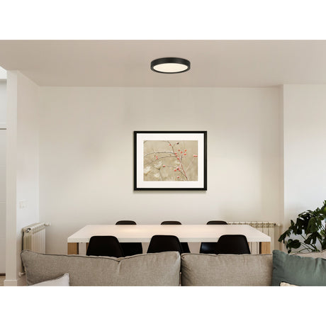 Idun-E round LED flush ceiling light in black with white diffuser, shown on a light ceiling above a dining area with framed botanical art and dark chairs.
