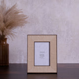 Rattan effect 4x6" photo frame with stand in natural woven texture and dark brown edging, shown on a dark wood surface against a neutral wall.