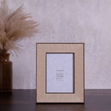 Rattan Effect Recycled Photo Frame with Stand in natural, holding a 5 x 7" insert, shown on a dark wood surface against a grey textured backdrop with dried pampas grass.