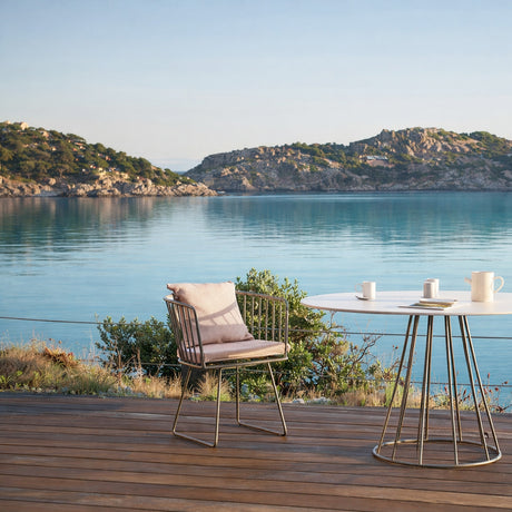 Illa aluminium garden dining armchair with slatted rod back, sled base, and beige cushions, shown on a wooden deck beside an oval outdoor table overlooking the sea.