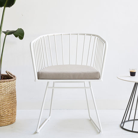 Illa aluminium garden dining armchair in white, with a rounded wire-rod back and arms, sled-style legs, and a beige seat cushion, shown indoors near a plant and side table.