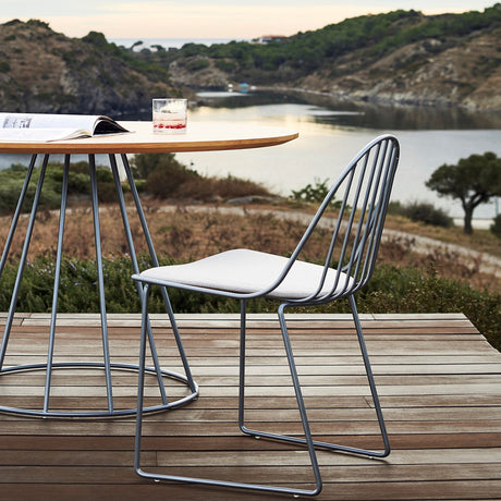 Illa Steel Stacking Garden Dining Chair in grey with slim rod back and sled base, shown with a round outdoor table overlooking a coastal landscape.