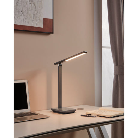 Iniesta LED Touch Table Lamp in grey and black, with slim bar-shaped head on a hinged upright and square base, shown lit on a desk beside a laptop and notebook.