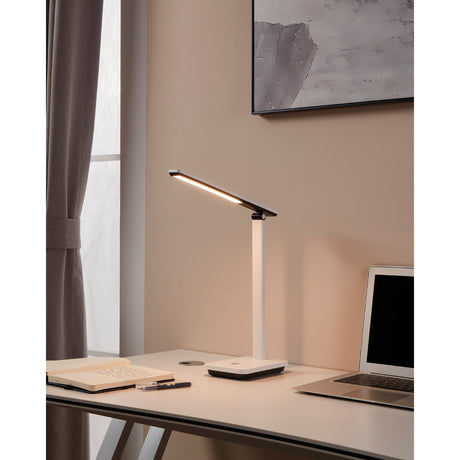 Iniesta LED touch table lamp with white stem and slim black-and-white bar head, shown lit on a desk beside a laptop and notebook.