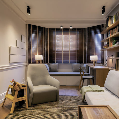 A cozy living room with modern decor includes a beige armchair, white sofa, and wooden chair. Wooden blinds and two small lamps accentuate the patterned rug. Shelves with books and a pink vase enhance the spaces charm, highlighted by a Focus Single Surface-Mounted Spotlight in black for adjustable lighting.