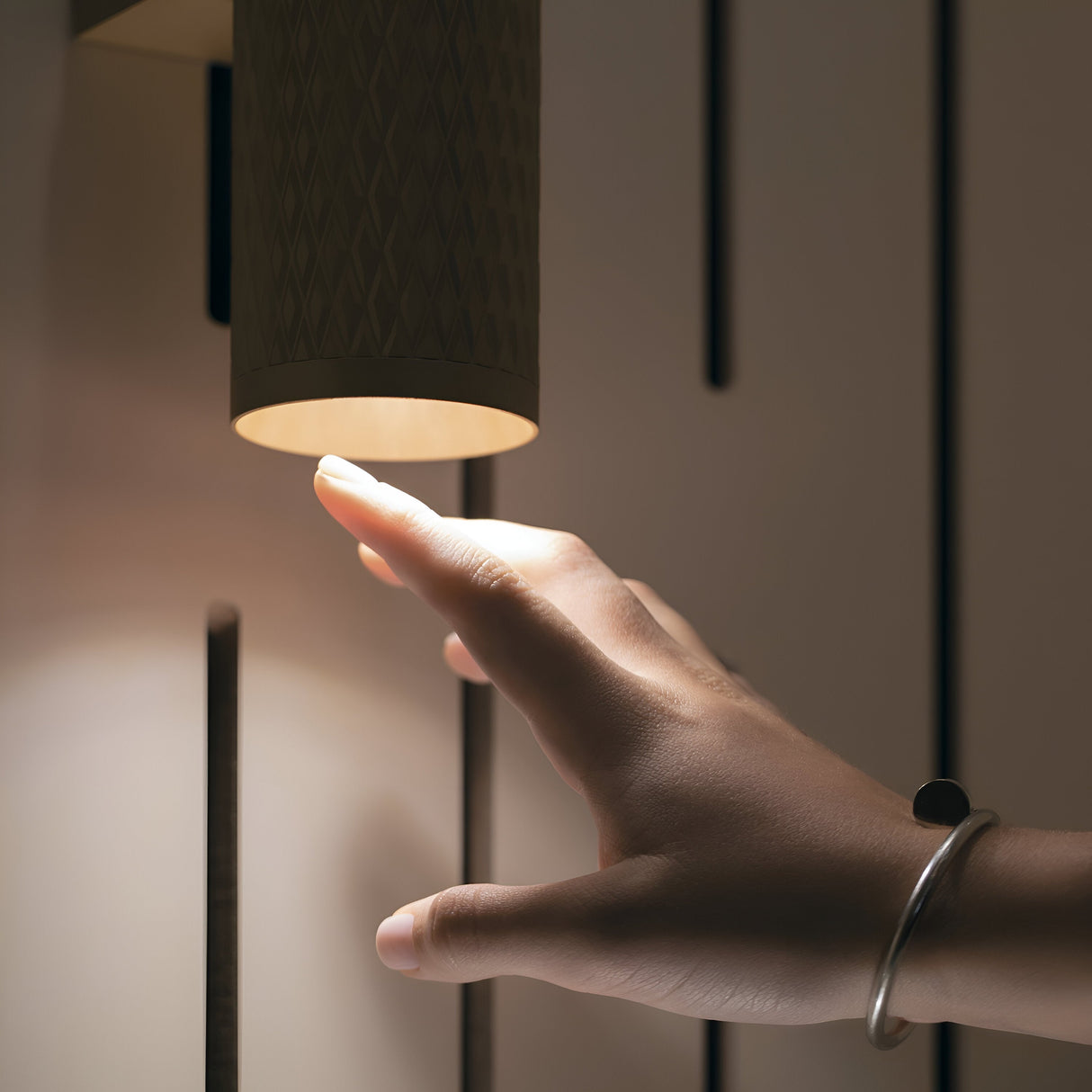 A hand reaches under the Focus Design Up & Down Wall Light - Black, casting a warm glow with its minimalist cylindrical design. Vertical dark slits on a light wall enhance the modern ambiance, while the wrist sports a simple bracelet with a small round charm.