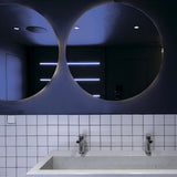 A modern bathroom showcases two circular mirrors reflecting a dim ambiance. The reflection includes the Atom Single Tilt Round Recessed Downlight in black, featuring an adjustable tilt. A white tiled backsplash and a rectangular sink with dual faucets adorn the counter.