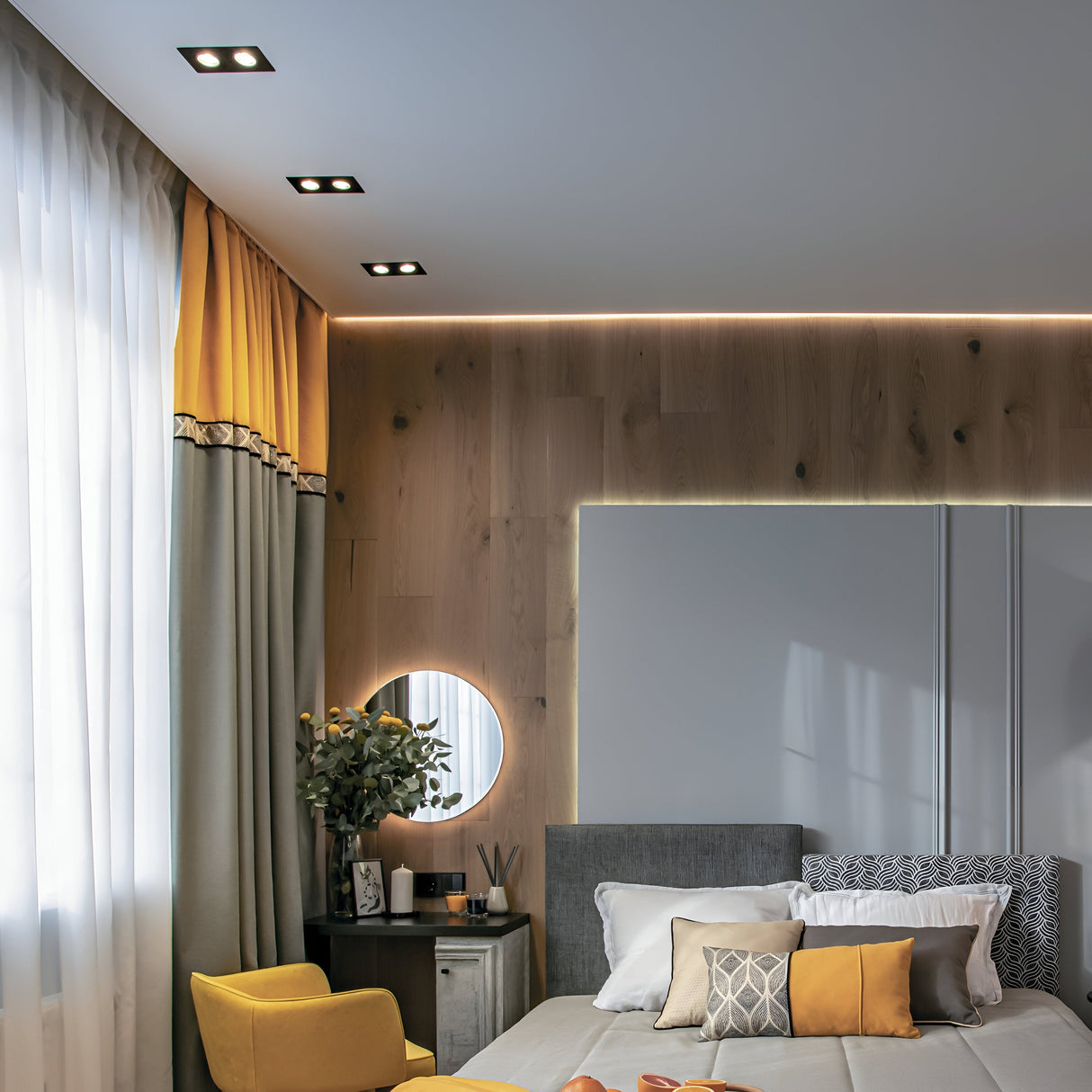 A modern bedroom features a neatly made bed with pillows in white, gray, and mustard. A side table sits beneath a round mirror adorned with plants and décor. Gray and yellow curtains frame the window, accented by an Atom Twin Tilt Rectangular Recessed Downlight - Black for perfect ambiance.