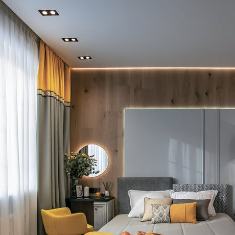 A modern bedroom features a neatly made bed with pillows in white, gray, and mustard. A side table sits beneath a round mirror adorned with plants and décor. Gray and yellow curtains frame the window, accented by an Atom Twin Tilt Rectangular Recessed Downlight - Black for perfect ambiance.