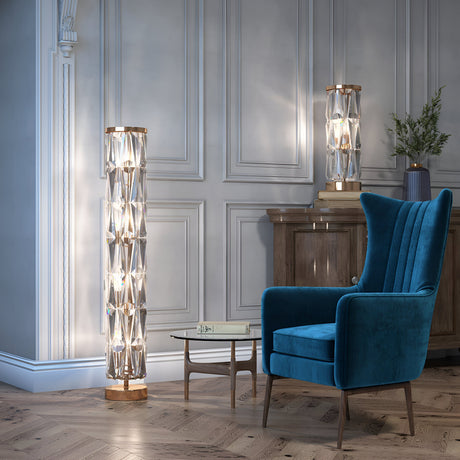 A room with a blue velvet armchair, a glass coffee table, and two Puntes Floor Lamps in gold and crystal featuring geometric designs. The light gray paneled walls complement a wooden cabinet with a vase of green foliage, while the herringbone floor enhances the luxurious lighting ambiance.