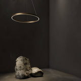 A minimalist room with a dimly lit, dark gray backdrop features the height-adjustable Rim LED Circular Suspension Light 60cm 4000K in Brass. Two large, irregularly shaped rocks rest on the floor, casting soft shadows.