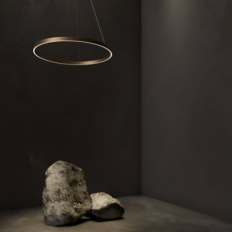 A minimalist room with a dimly lit, dark gray backdrop features the height-adjustable Rim LED Circular Suspension Light 60cm 4000K in Brass. Two large, irregularly shaped rocks rest on the floor, casting soft shadows.