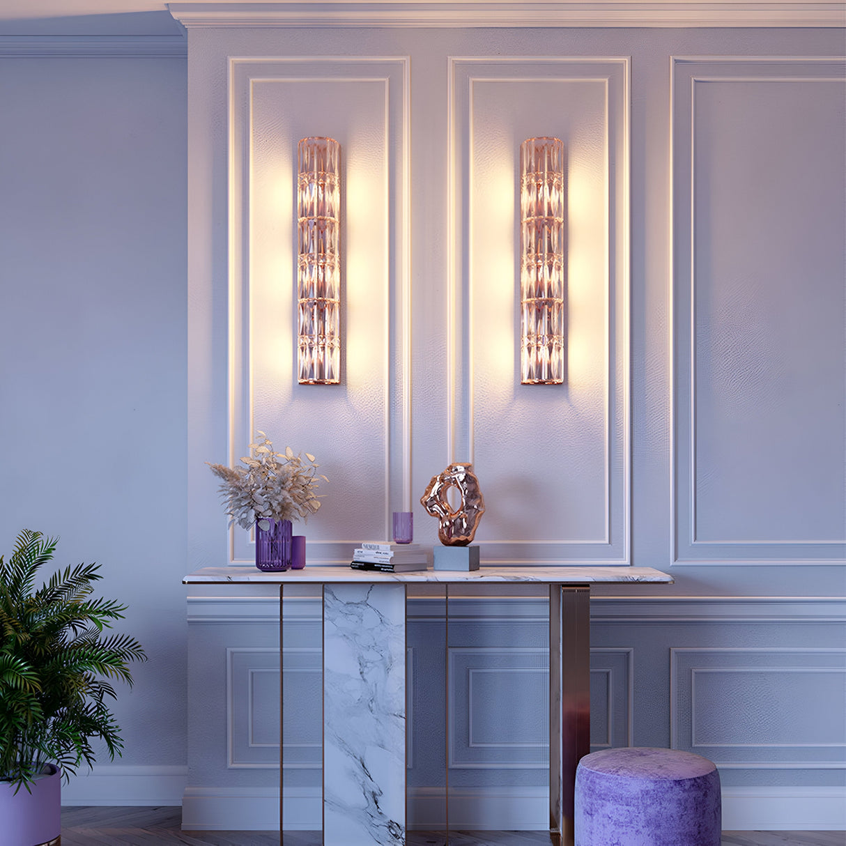 An elegant interior features the Facet 3 Light Long Wall Light in gold, casting warm light across the paneled wall. A marble console table with a flower vase and geometric sculpture sits below. A purple ottoman and a potted plant complete the scene.