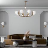 A modern living room showcasing a brown sectional and two small white side tables. A black coffee table holds a white vase with dried plants, while the Rendez-vous 8 Light Chandelier - Brass and matching wall sconces illuminate the space against light gray walls.