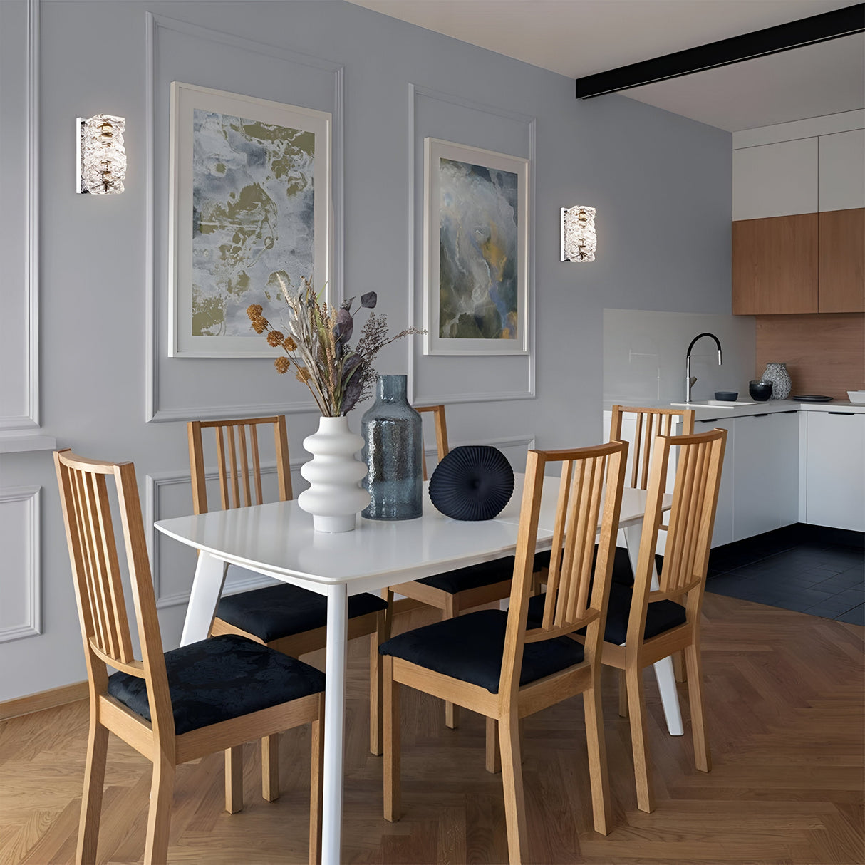 A contemporary dining area includes a white table and six wooden chairs with black cushions. Two abstract paintings complement the Coil LED Wall Light - Chrome on the gray wall. A vase with dried flowers sits on the table, with chrome finish elements enhancing the kitchen in the background.