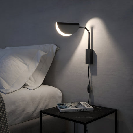 A modern bedroom has a sleek Mollis Wall Light - Black, casting LED lighting on a gray wall. Below, a small black table with a book and magazine sits beside a bed adorned with white pillows and linens, enhancing the cozy minimalist design.