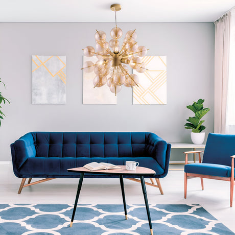 In a modern living room, a blue velvet sofa, matching chair, and geometric rug set the scene. A wooden coffee table holds a book and mug, abstract art adorns the gray wall, and the Bolla 6 Light Chandelier in brass with amber glass shades casts an elegant glow. A potted plant is in the corner.