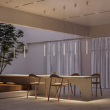 A modern dining area features a minimalist design with a sleek table and three wooden chairs. The Pro Base LED Thin Pendant Light in white stylishly hangs above, casting a gentle glow. Soft natural light filters through sheer curtains, while a tall plant adds greenery to the serene space.