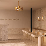 A minimalist interior in beige tones features modern chairs against a stone wall, highlighted by the Tropic 6 Light Chandelier in brass overhead. Textured surfaces add depth, and a partially visible motivational quote reads LIFE IS INSPIRATION.