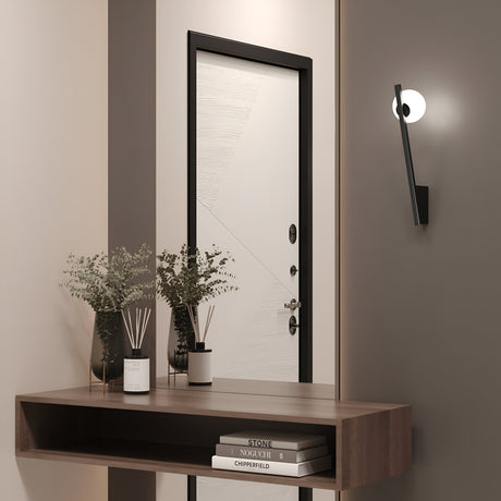 A minimalist hallway features a wooden console table with books, two plants, and a decorative diffuser. The Kazimir Wall Light - Black With White Opal Glass illuminates the space, accentuating the geometric-designed door and evoking an avant-garde artistry with minimalistic silhouettes.