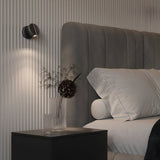 A modern bedroom features a gray upholstered headboard, white bedding, and a black nightstand. The nightstand holds two vases—one with dried flowers and one empty. Soft lighting is provided by the minimalist Nuance LED Movable Head Wall Light in black, offering energy-efficient illumination.